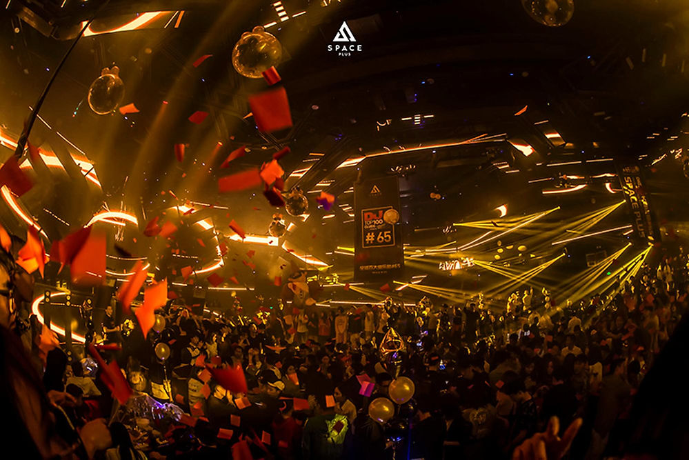 Crowded nightclub with red confetti and golden lights. Energetic atmosphere, vibrant beams, and "Space Plus #65" banner visible.