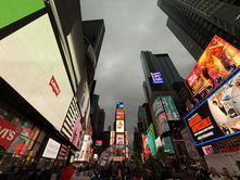Times Square at dusk, crowded with people under towering buildings. Bright billboards display vibrant ads and text, creating a lively mood.