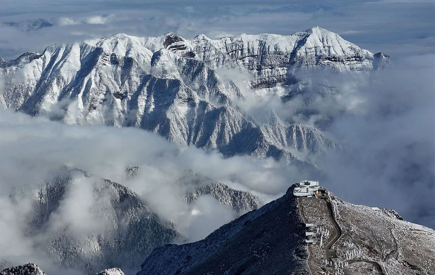 Snowy mountain peaks with a building on a ridge. Clouds surround the mountains, creating a serene and majestic winter landscape.