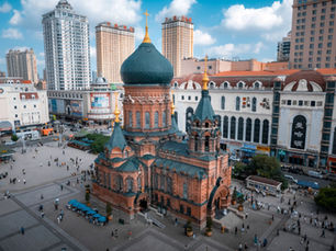Aerial view of Saint Sophia Cathedral with green domes in a bustling square. Surrounded by modern buildings under a bright blue sky.