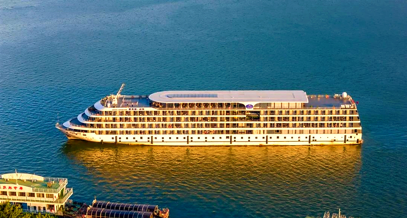 Large white cruise ship on calm blue water, sunlit with a modern design. Nearby a smaller boat with Chinese text. Peaceful atmosphere.