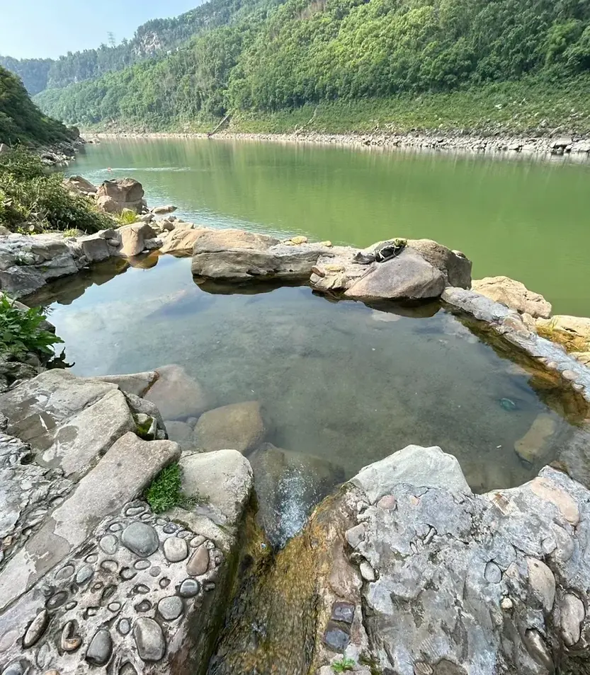 Natural rock pool by a calm, green river surrounded by lush forested hills. The rocky foreground has pebbles and grass, creating a serene scene.