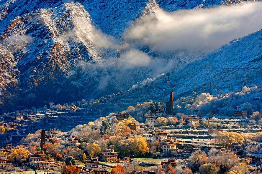 Snow-covered village at mountain base with colorful autumn trees. Misty clouds envelop the blue-tinted peaks, creating a serene atmosphere.