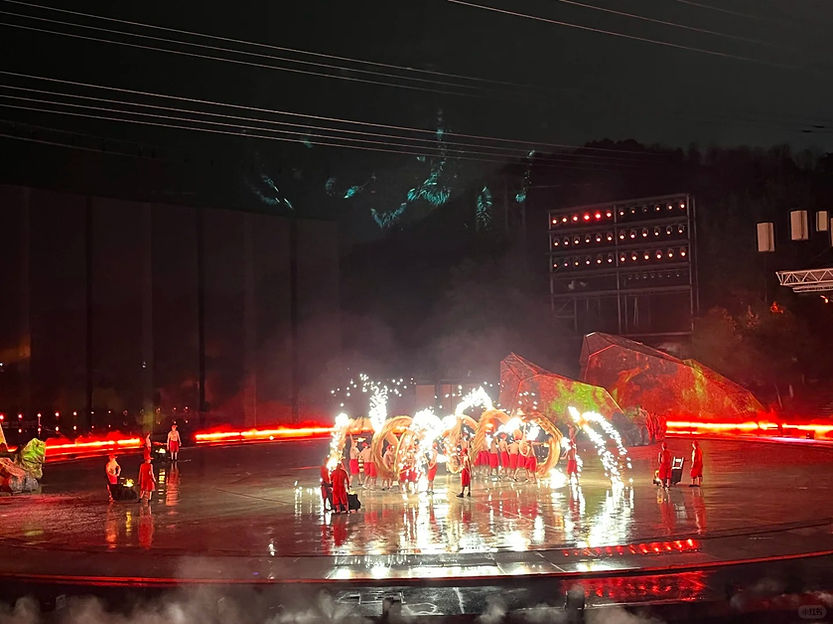 Performers in red costumes on a wet stage with fire effects and sparklers in a dark setting. Bright red lights highlight the scene.