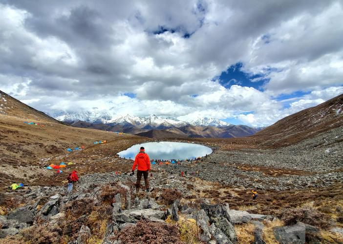 A person in a red jacket stands by a mountain lake surrounded by snow-capped peaks and tents under a cloudy sky. Rugged terrain.
