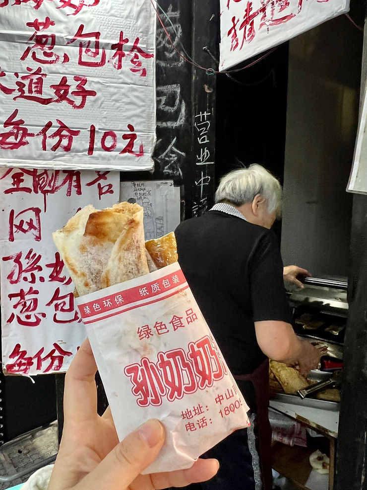 Hand holding a filled pastry in a paper bag with red text near a street vendor with white hair cooking. Chinese signs visible in the background.