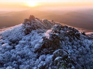 Snow-covered mountain peaks at sunrise, bathed in warm orange light. Frosty trees surround the rocks, creating a serene winter landscape.