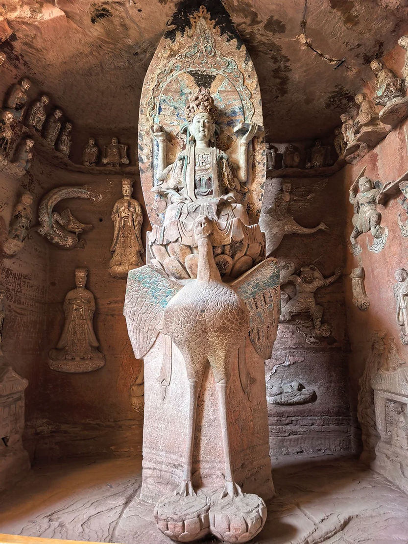Stone statue of a seated figure on a lotus in a cave, surrounded by intricate carvings and smaller statues, with earthy tones and textures.