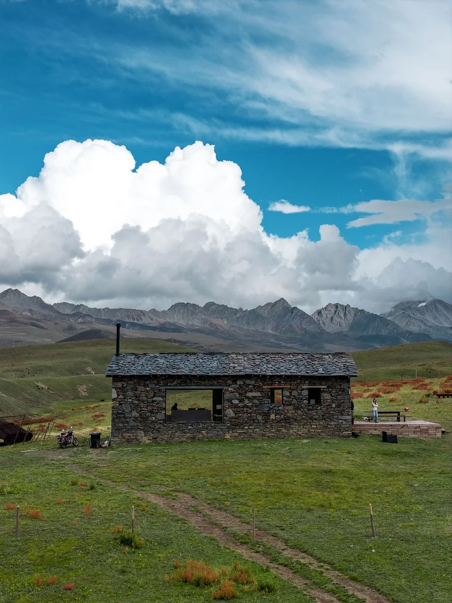 Stone house on a grassy field with a motorcycle and person nearby, mountains in the background under a bright blue sky with fluffy clouds.