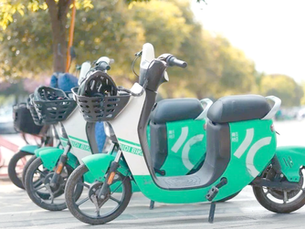 Three green electric scooters with helmets in baskets are parked on a sunny sidewalk. Background shows trees and a blurred park.
