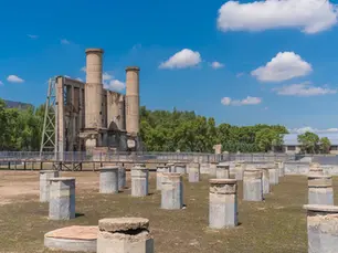 Ruins of an old industrial building with two tall smokestacks in a grassy field, surrounded by short concrete pillars. Clear blue sky above.