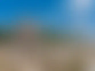 Ruins of an old industrial building with two tall smokestacks in a grassy field, surrounded by short concrete pillars. Clear blue sky above.