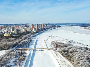 Aerial view of a snow-covered city beside a frozen river. Buildings and trees line the riverbank. The sky is clear and blue.