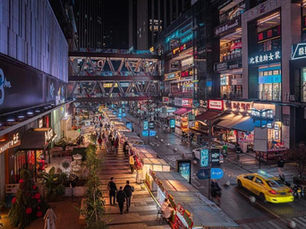 Night market scene with busy street, colorful shops, and illuminated signs. People walk along stalls; a yellow taxi passes by, vibrant atmosphere.