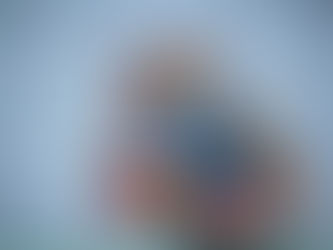 A person in traditional attire stands before a colorful deity painting on a white wall, surrounded by offerings and a lantern.