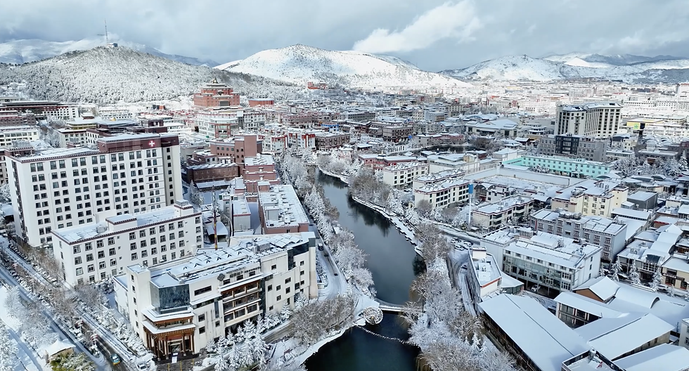 Snow-covered cityscape with buildings lining a winding river, framed by snowy hills under a cloudy sky. Buildings mostly white with red accents.
