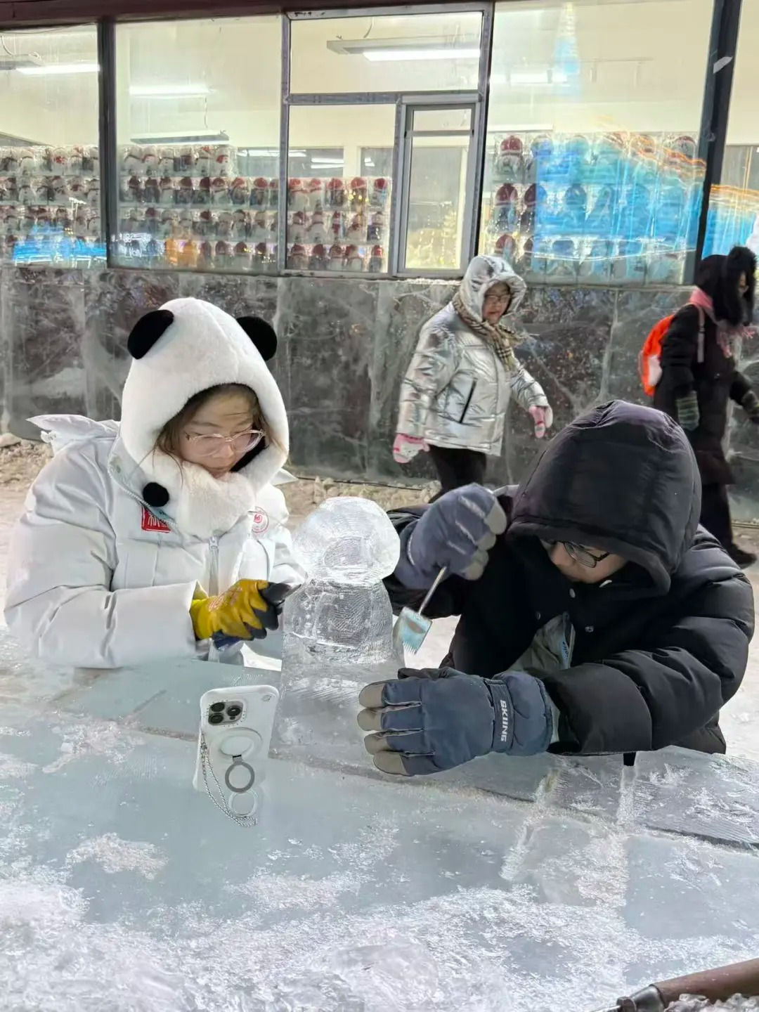 Two people in winter clothes carve an ice sculpture on a snowy table. A phone stands nearby. Others pass by in the background.