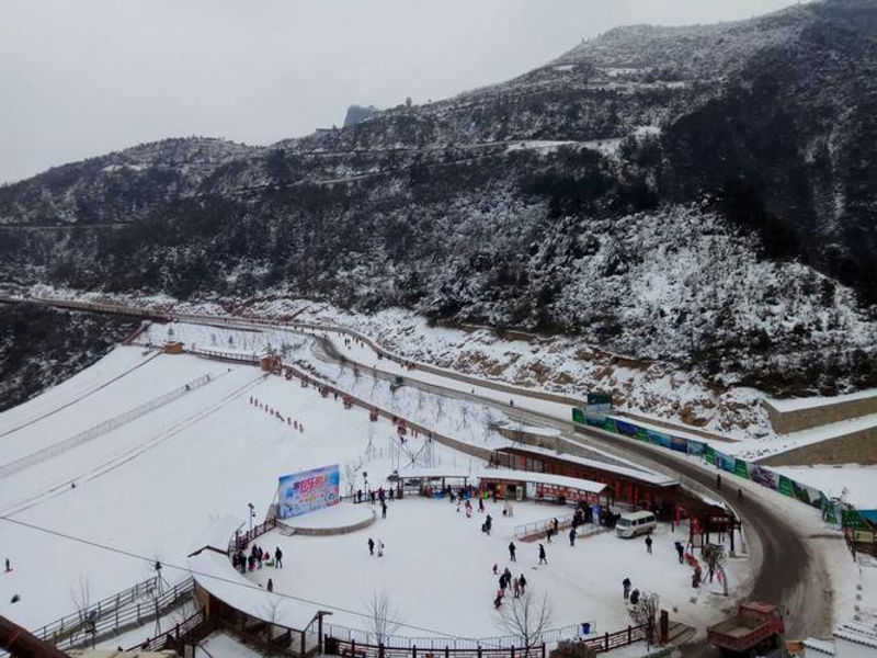 People ski on a snowy slope set against a backdrop of snow-covered mountains. A banner and colorful buildings add vibrancy to the scene.