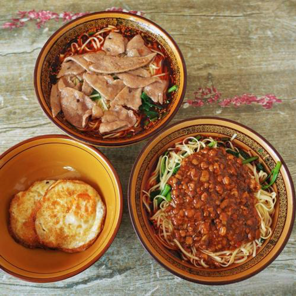 Bowls of noodles with beef slices and meat sauce, and a yellow bowl with two fried eggs on a rustic wooden table with floral accents.