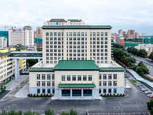 Large beige building with green roofs, set in an urban landscape. Text in red above entrance. Overcast sky and nearby cranes visible.