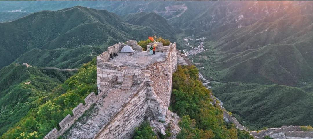 Person stands on ancient stone tower with tent, surrounded by green mountains, capturing a scenic view at sunrise; a red flag is visible.