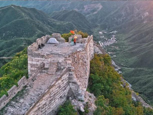 Person stands on ancient stone tower with tent, surrounded by green mountains, capturing a scenic view at sunrise; a red flag is visible.