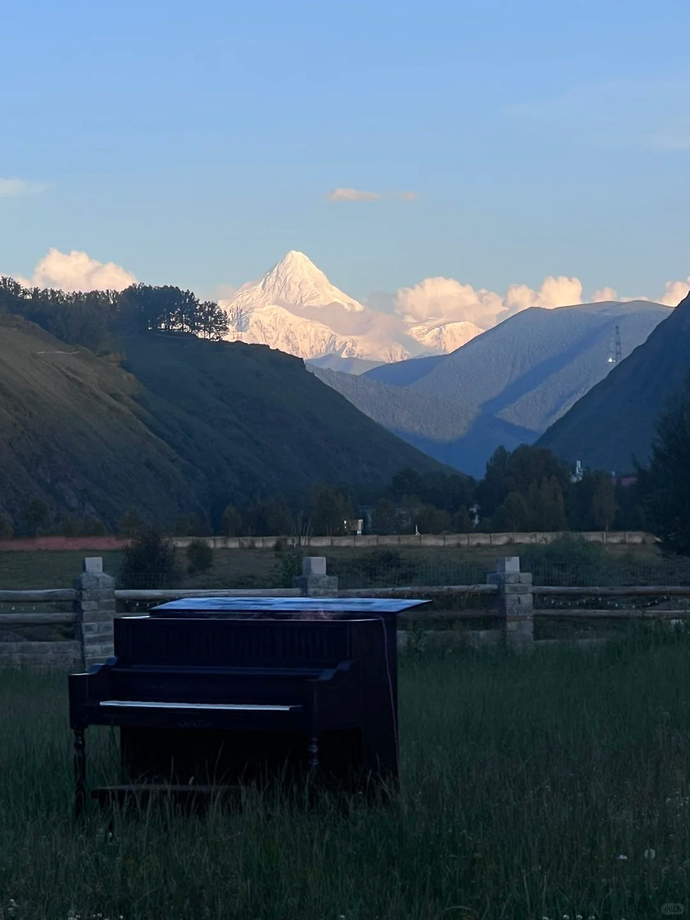 A black piano sits in a grassy field with mountains and a snow-capped peak in the background under a blue sky, creating a serene scene.
