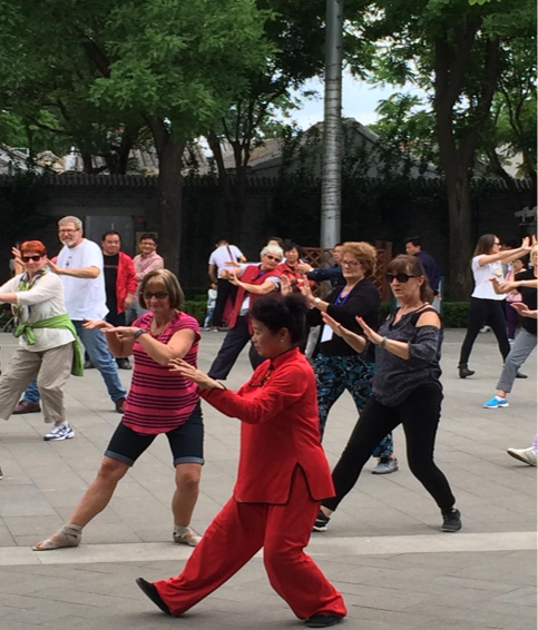 People practicing Tai Chi in a park with green trees, wearing colorful outfits. The setting is calm and focused, with no visible text.
