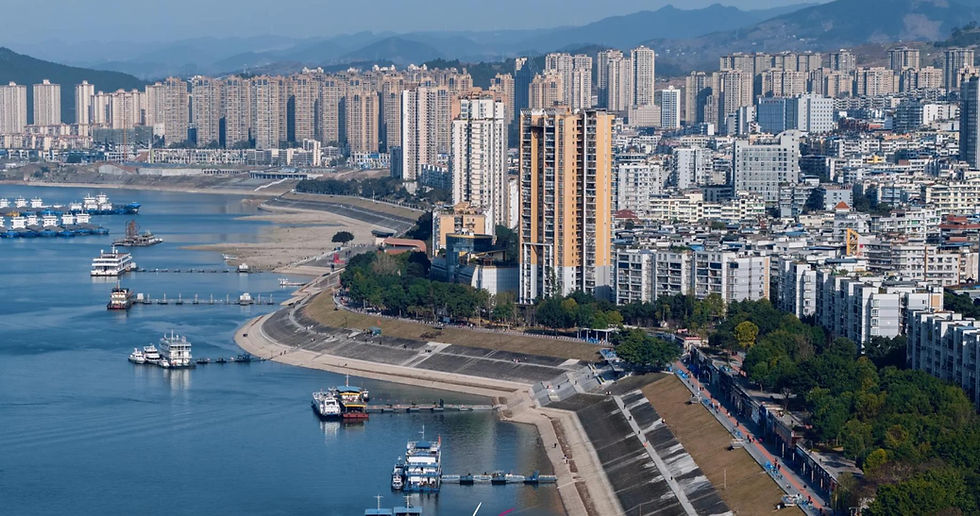 Cityscape with tall buildings lining a river, boats docked along the shore. Blue water, clear sky, and mountains in the distant background.