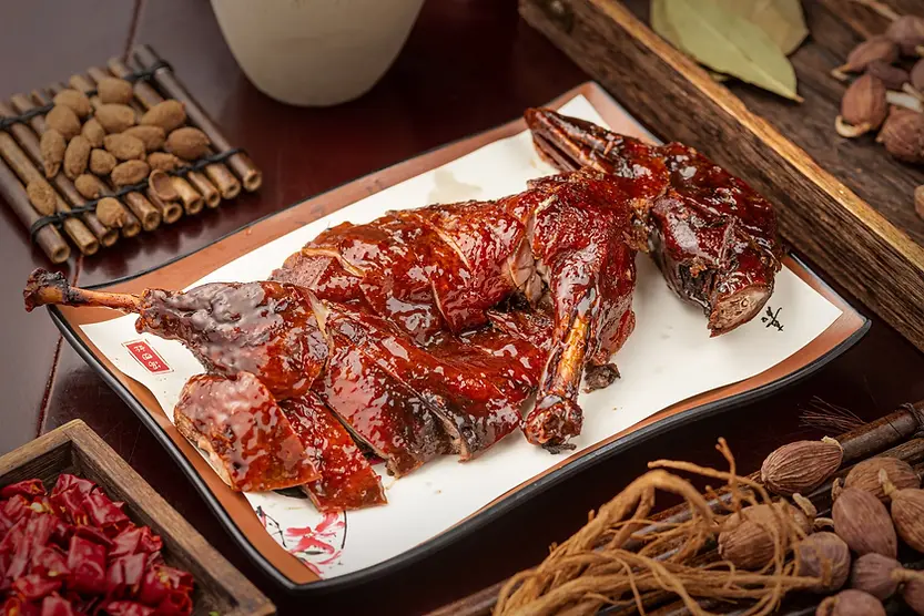 Roasted duck on a white plate, glazed and shiny, surrounded by spices on a wooden table. Rich brown tones and a savory mood.