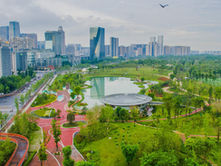 Aerial view of a green park with winding paths, a circular building by a pond, a bird in flight, and a city skyline in the background.