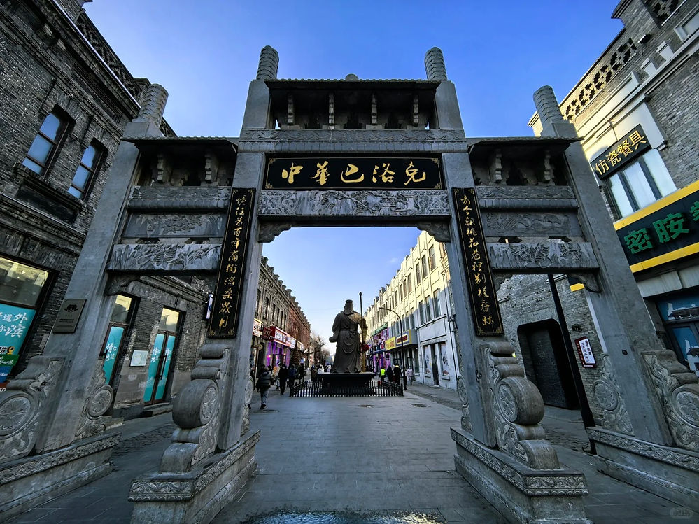Stone archway with Chinese text, flanked by traditional buildings under a clear blue sky. People walk below, and a statue stands in the center.
