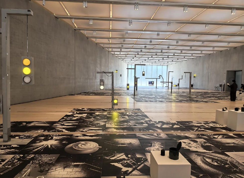 Gallery with traffic lights on a patterned floor, featuring black and white art. Two people observe the minimalist setting.