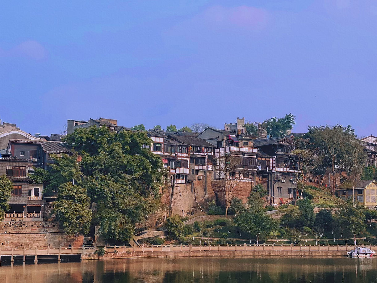Historic hillside village with traditional buildings and lush trees, reflected in calm river under a clear blue sky. Warm, serene atmosphere.