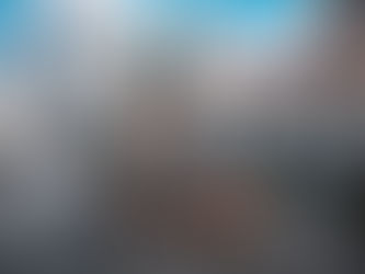 Red-brick church with green domes in a busy city square. People walk nearby. Tall buildings surround under a bright blue sky.