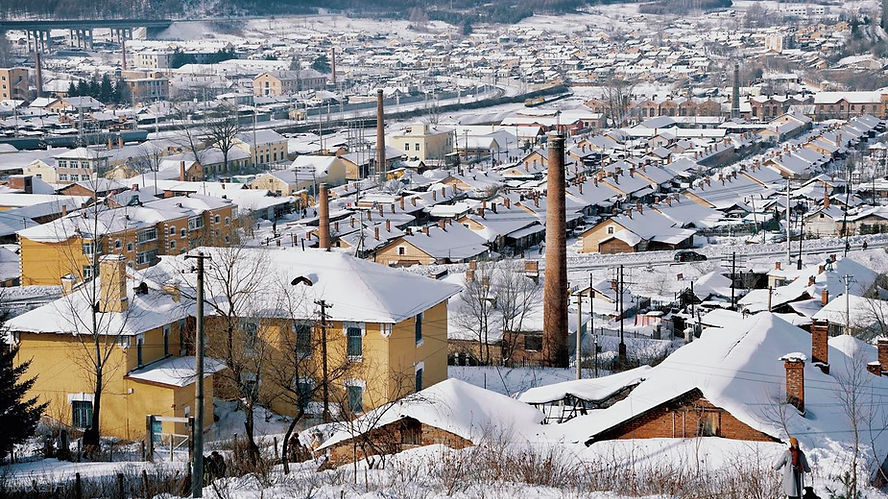 Snow-covered town with yellow buildings and chimneys, set against a backdrop of snowy hills and a distant bridge under a clear sky.