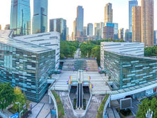 Modern cityscape with tall, reflective skyscrapers surrounded by lush greenery. A central pathway leads through a plaza with colorful decor.