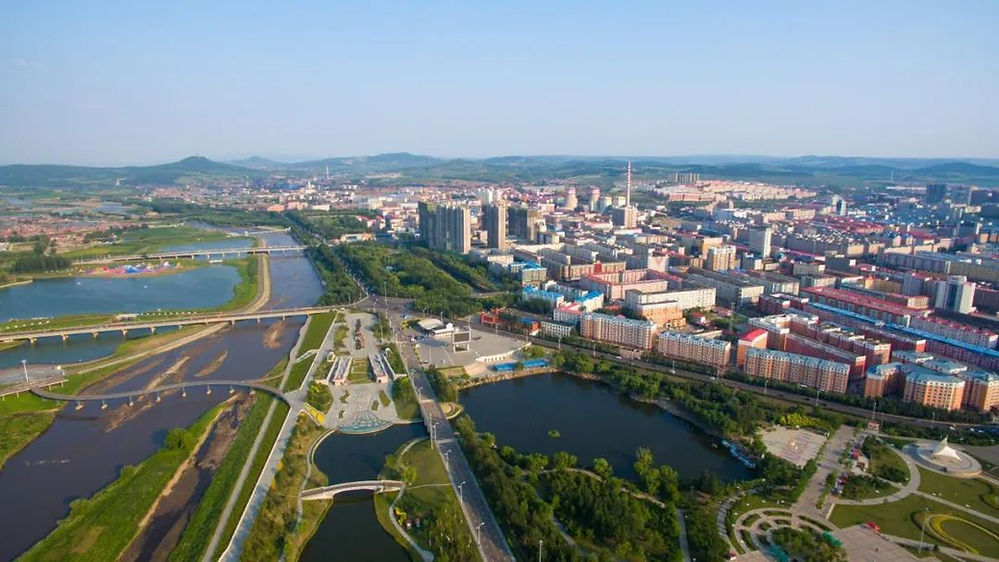 Aerial view of a city with colorful buildings, surrounded by rivers and green spaces. Clear sky and distant hills in the background.