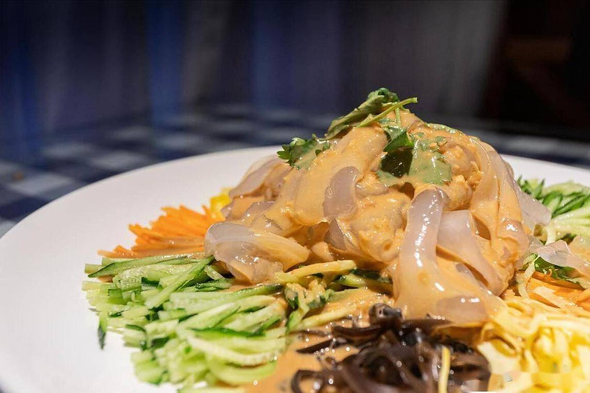 A plate of vibrant salad with sliced cucumbers, shredded carrots, and noodles topped with creamy peanut sauce, set on a blue checkered table.