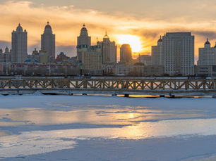 Frozen river with icy surface, wooden railing bridge in foreground, city skyline at sunset in the background, warm glow in the sky.