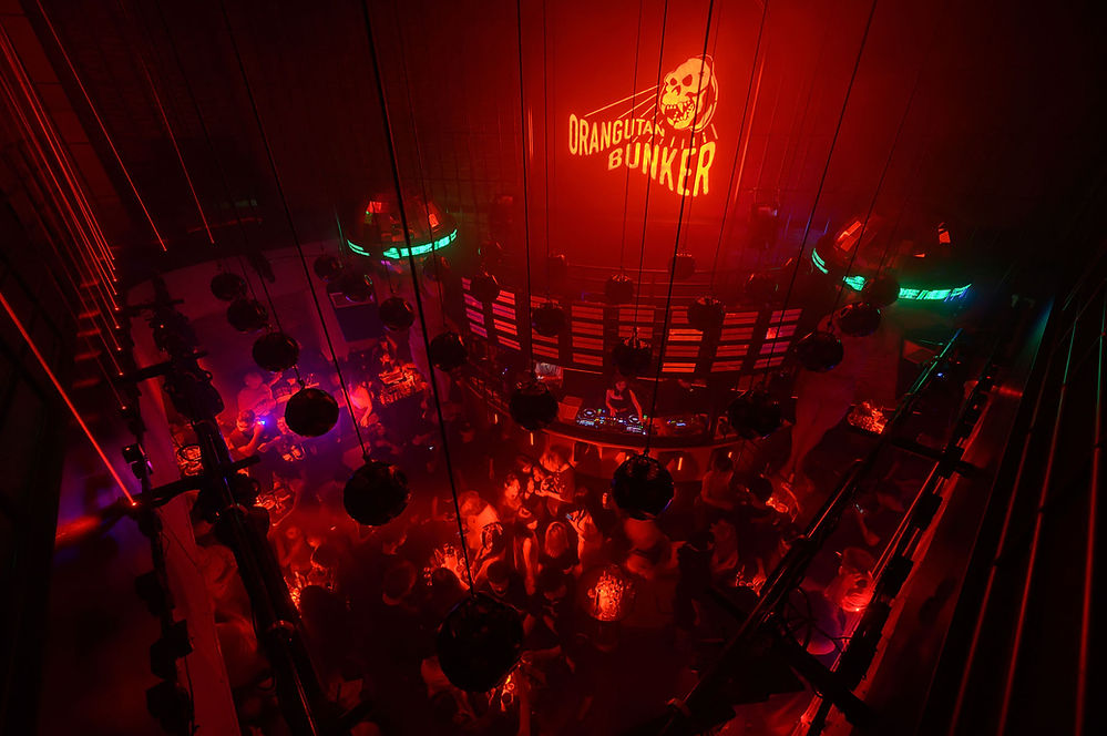 Nightclub scene with people dancing under red and green lights. DJ at the booth. "Orangutan Bunker" logo glows on the wall. Energetic mood.