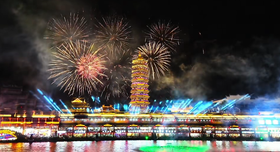 Fireworks burst in the night sky above an illuminated pagoda and buildings, with colorful reflections on the water, creating a festive scene.