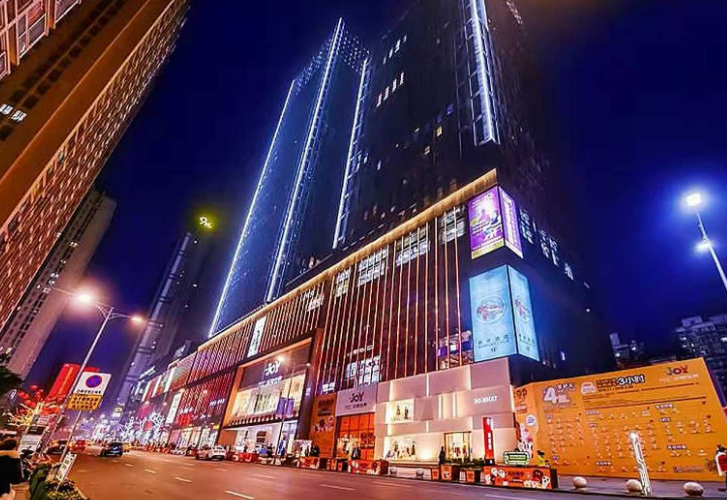Tall, illuminated buildings along a busy city street at night. Bright signage displays various advertisements. Streetlights cast a warm glow.