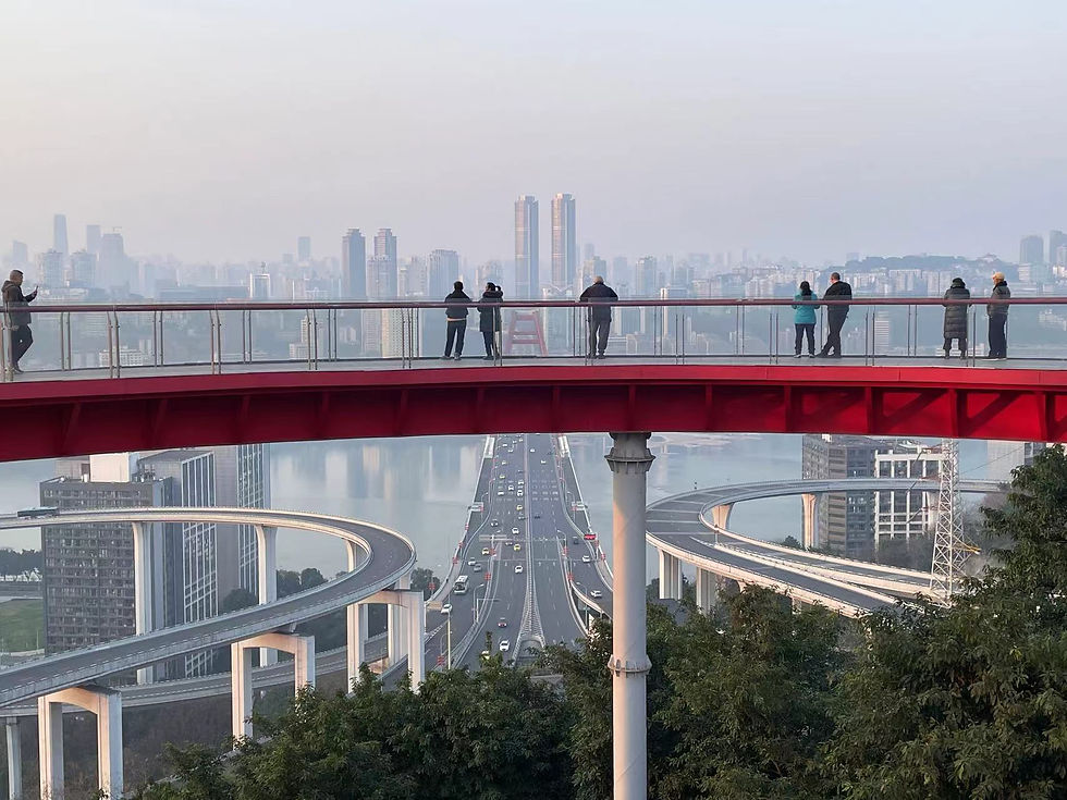 People stand on a red bridge overlooking a cityscape with tall buildings. A highway curves below. Sky is clear, creating a calm mood.