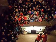 Crowd gathered in a dimly lit area watching two performers in red creating shadow puppets on a lit screen. The mood is curious and engaged.
