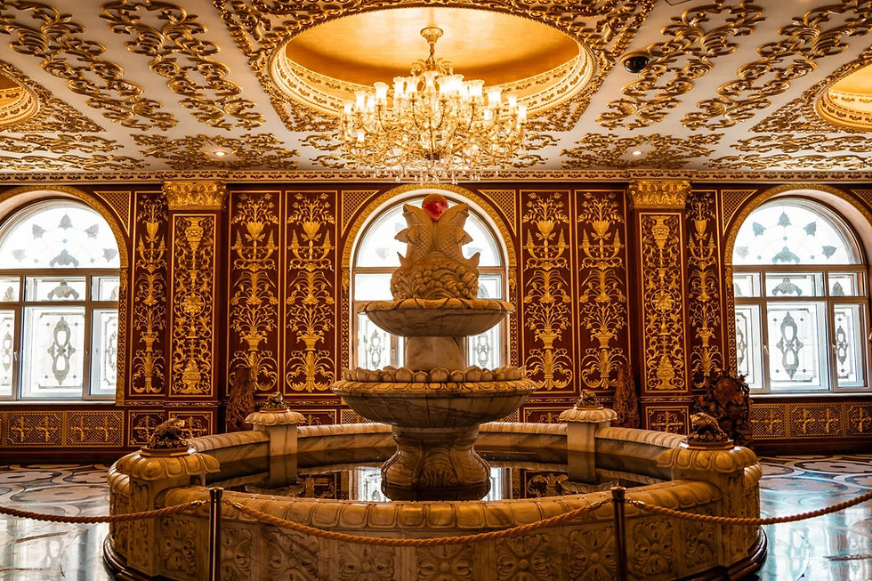 Ornate room with a central marble fountain. Gold-patterned walls, chandelier, and arched windows create a lavish, regal atmosphere.