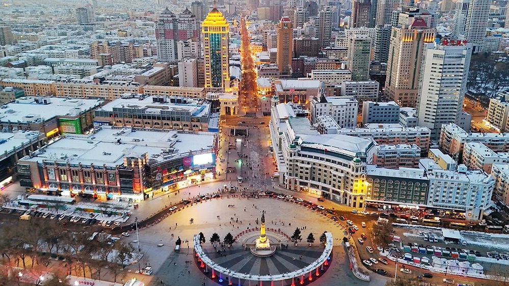 Aerial view of a vibrant cityscape at dusk with snow-dusted buildings, bright lights, a central circular plaza, and light trails from cars.