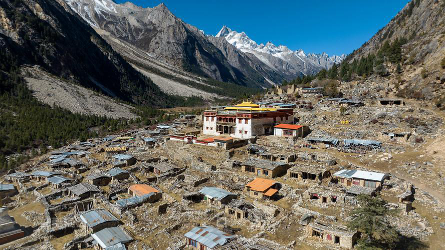 Mountain village with stone houses and a colorful monastery under a clear blue sky. Snow-capped peaks in the background, creating a serene scene.