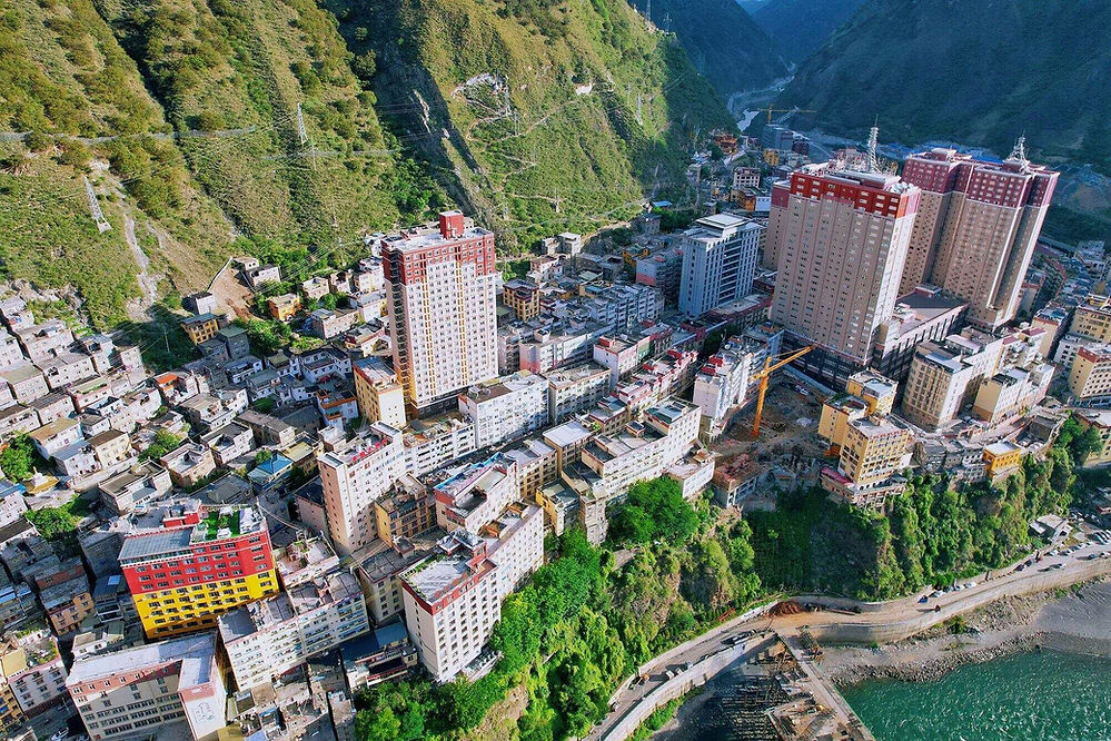 Aerial view of a colorful city with tall buildings near lush, green mountains and a river. A crane indicates ongoing construction.