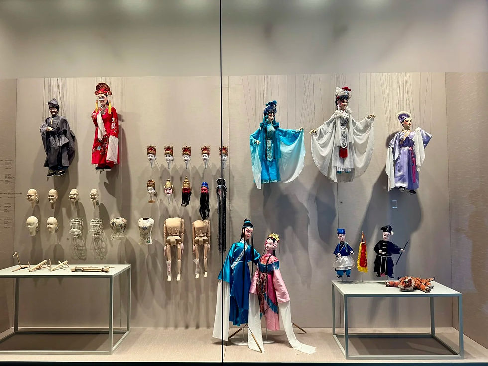 Colorful puppets in traditional attire are displayed on a wall alongside puppet parts. Two tables hold accessories in a museum setting.